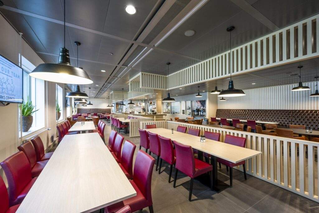 Modern bedrijfsrestaurant aan het Koningin Wilhelminaplein met lange houten tafels, rode stoelen en industriële plafondlampen.